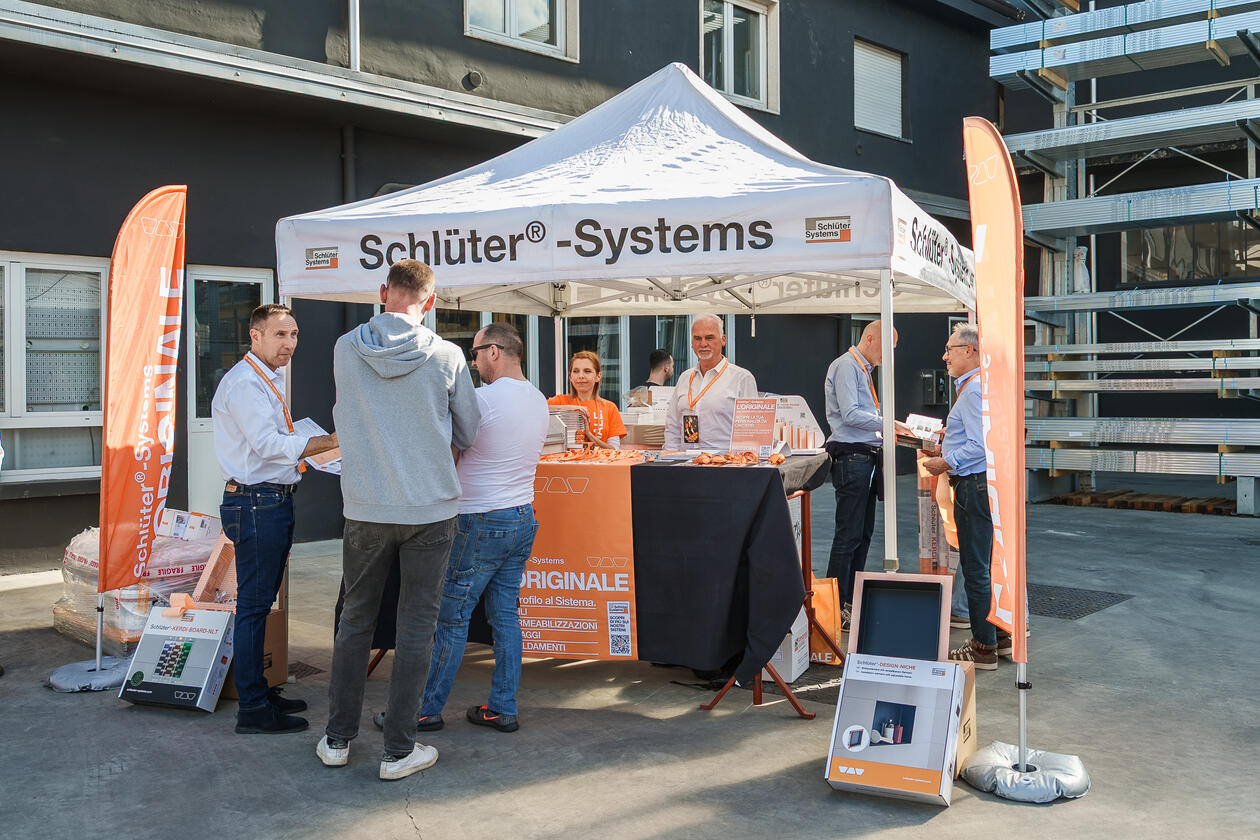 La foto mostra uno stand fieristico di Schlüter-Systems con i relativi prodotti. Sotto e davanti al padiglione ci sono in totale otto persone.