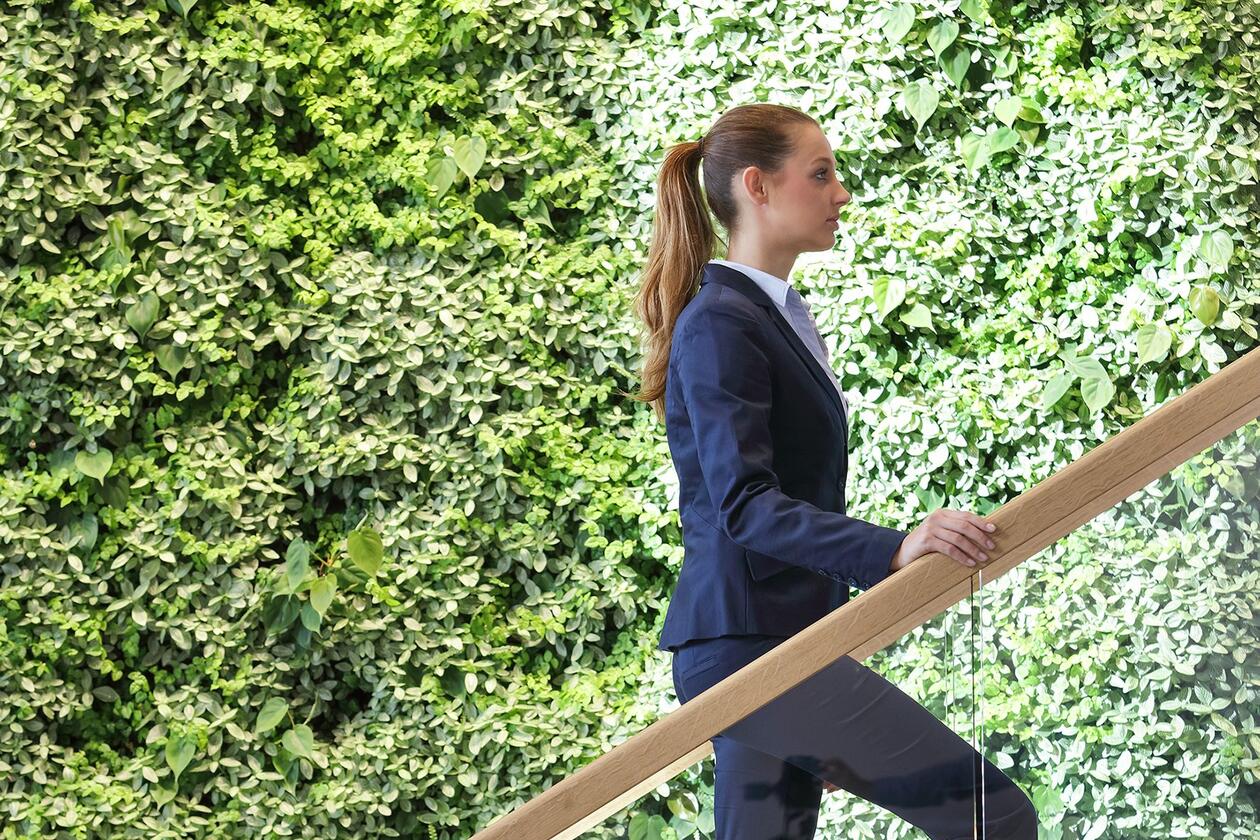 Ansicht einer Frau, die vor einer grünen Blätterwand eine Treppe hochsteigt.
