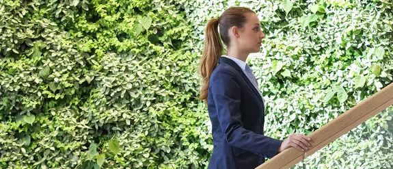Mladá žena stoupá po schodech, v pozadí je zelená stěna porostlá rostlinami.