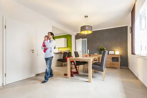 A father wearing socks walks through the dining room with a baby in his arms.