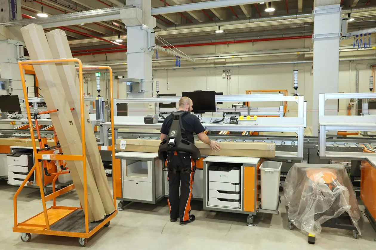 Male Schlüter-Systems employee lifts goods with the aid of an exoskeleton in front of a packaging system.