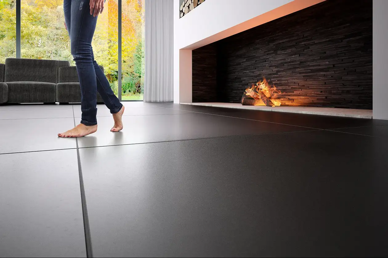 The legs of a woman can be seen walking barefoot across a floor laid with large, dark tiles in a modern living room in front of a wide fireplace.