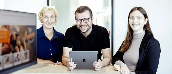 Three Schlüter-Systems employees standing at a table, the person in the middle is holding a tablet computer in his hands.