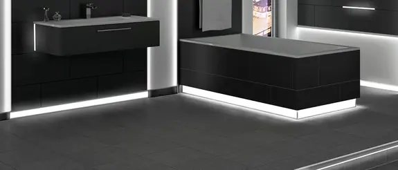Floor area of a black-tiled bathroom with illuminated skirting profiles on the wall and edge of the bathtub