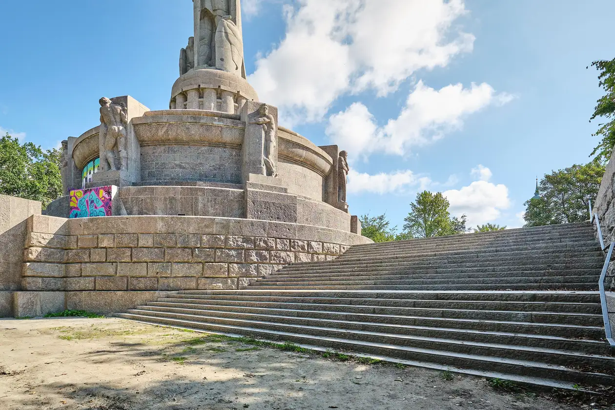 Ein breiter Treppenaufgang führt zum Bismarck-Denkmal auf einer Anhöhe. Im Hintergrund sind einige Baumkronen und blauer Himmel mit vereinzelten Wolken.