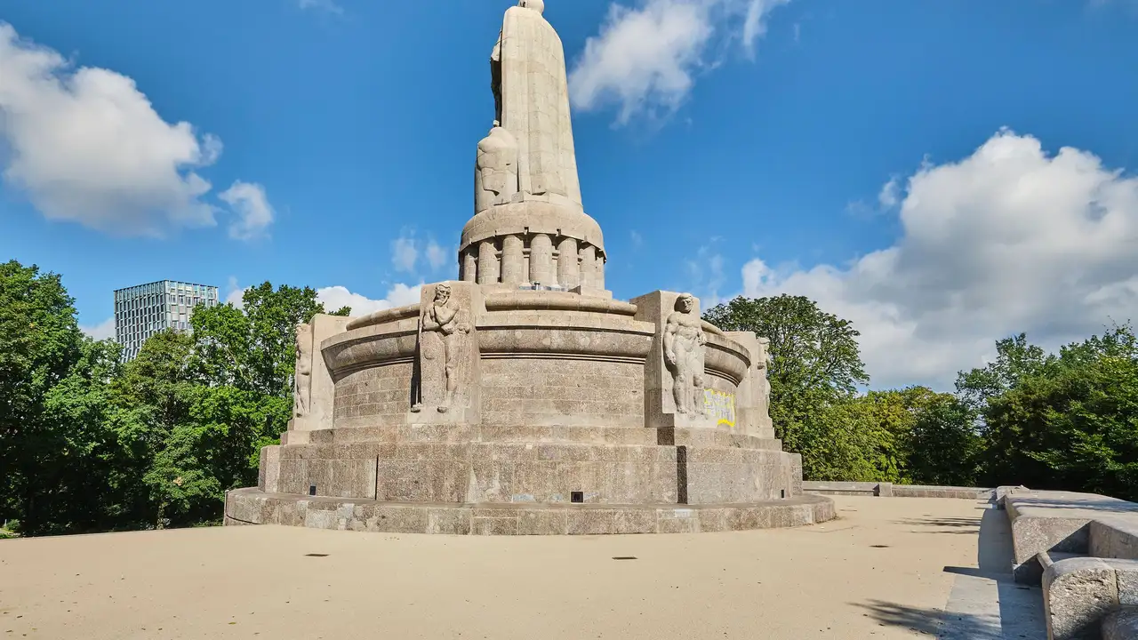 Das Bismarck-Denkmal auf einem großen Platz, es ist der Rücken der Figur zu sehen vor einem blauen Himmel mit einigen Wolken und grünen Baumkronen.