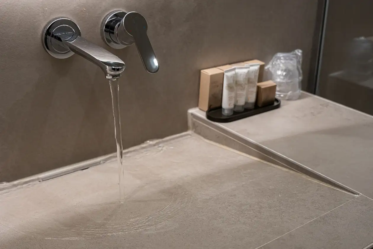 Silberne Armatur mit fließendem Wasser über einem Waschtisch. Die verbauten Profile Schlüter-JOLLY und KERDI-LINE-VARIO in steingrauer Strukturbeschichtung fügen sich harmonisch in die beige Fliesenoptik ein. Auf dem Waschtisch sind einige Waschartikel. 