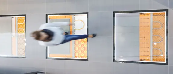 Frau läuft über Fliesenboden mit Sichtfenster auf die Fußbodenheizung BEKOTEC-THERM.