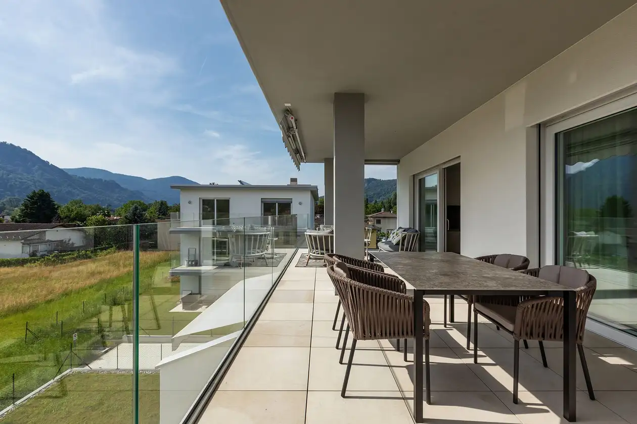 Hell gefliester Balkon mit gro&szlig;em Esstisch, im Hintergrund die Aussicht ins Gr&uuml;ne und auf Bergpanorama