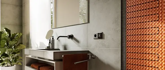 Ausschnitt eines Badezimmers mit Waschbecken, Handtuchhalter, Spiegel und unterhalb der Fliesen angelegter elektrischer Fl&auml;chenheizung an der Wand. Daneben befindet sich das Raumthermostat.
