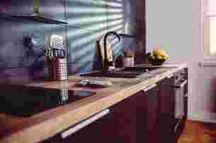 Moderne Küche mit dunklem Fliesenspiegel, Holzarbeitsplatte, schwarze Geräte, schwarze Armatur und zwei mattschwarzen Schlüter-SHELF Designablagen an der Wand. 