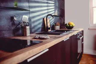 Moderne K&uuml;che mit dunklem Fliesenspiegel, Holzarbeitsplatte, schwarze Ger&auml;te, schwarze Armatur und zwei mattschwarzen Schl&uuml;ter-SHELF Designablagen an der Wand. 