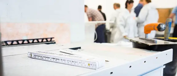 Ebene Arbeitsfläche in eimem Schulungsraum mit Profilkante, Zollstock, Kelle, Spachtel und Schulungsteilnehmern im Hintergrund