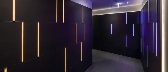 Blick in einen geschwungenen Gang mit schwarz gefliesten W&auml;nden und bunt leuchtenden Lichtlinien in den Fugen