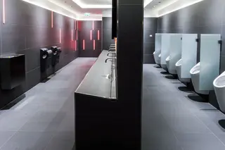 Sanit&auml;rbereich f&uuml;r Herren mit orange leuchtenden Lichtprofilen zwischen den dunklen Fliesen und einem gro&szlig;en Wasch- und WC-Bereich