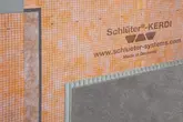 Wandfläche mit orangener KERDI-Abdichtungsbahn und eingebetteter Fliese