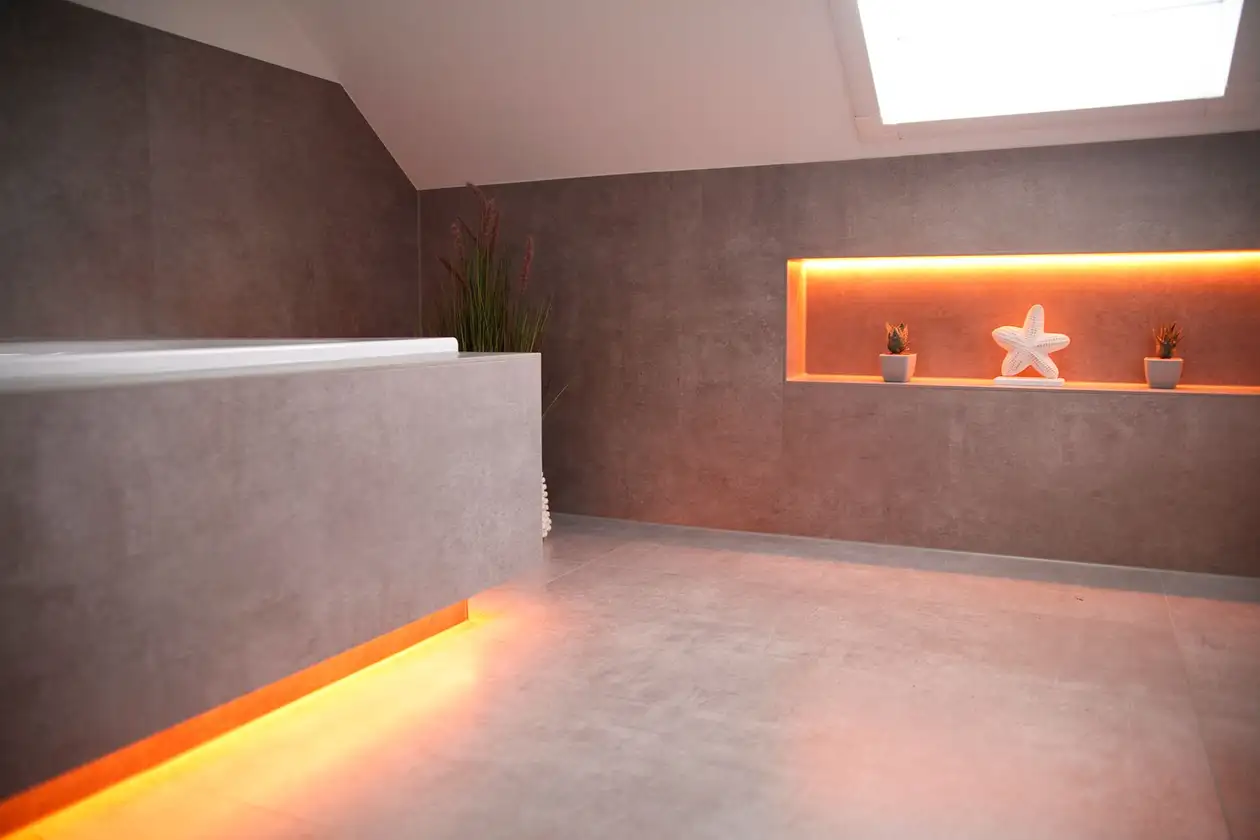 Ein beige-grau gefliestes Badezimmer mit Dachschr&auml;ge und warmer Beleuchtung an Badewanne und Nische