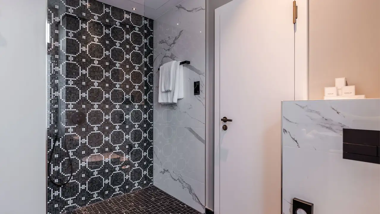 Cuarto de ba&ntilde;o terminado tras la renovaci&oacute;n con baldosas decorativas oscuras en la ducha y baldosas de m&aacute;rmol blanco en las paredes
