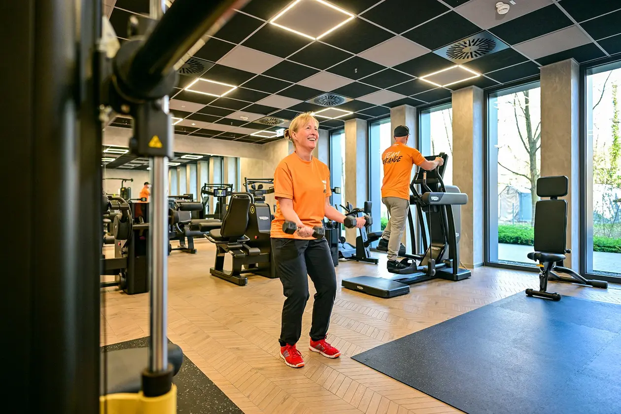 Une employ&eacute;e de Schl&uuml;ter-Systems soul&egrave;ve des halt&egrave;res dans une salle de sport, avec en arri&egrave;re-plan un employ&eacute; de Schl&uuml;ter-Systems sur un stepper
