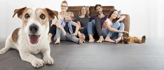 Une joyeuse famille de cinq personnes, un chien et un chat sont assis pieds nus sur un sol carrelé gris devant un canapé marron.
