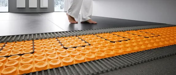 Une femme marche sur du carrelage. Devant, le chauffage au sol électronique Schlüter-DITRA-HEAT-E posé en dessous, vu en coupe.