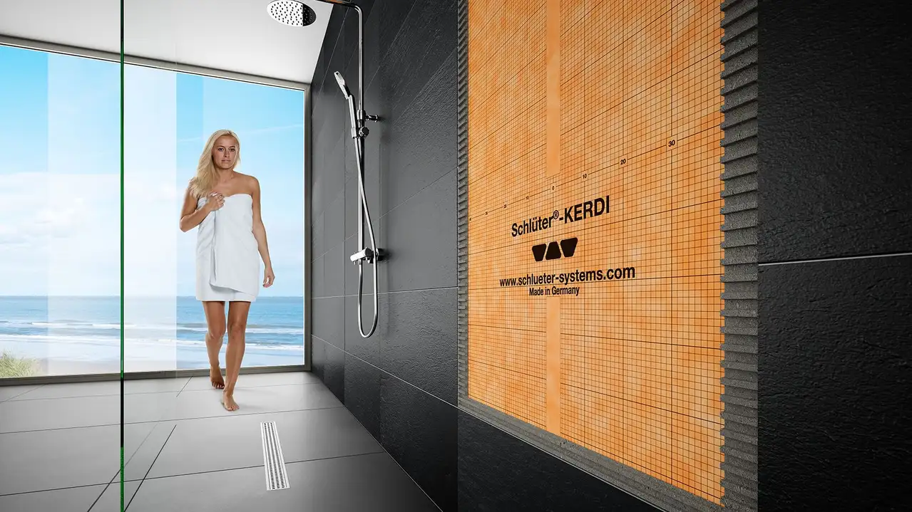 Une femme dans une salle de bains au carrelage sombre, un chauffage mural DITRA-HEAT-E en coupe