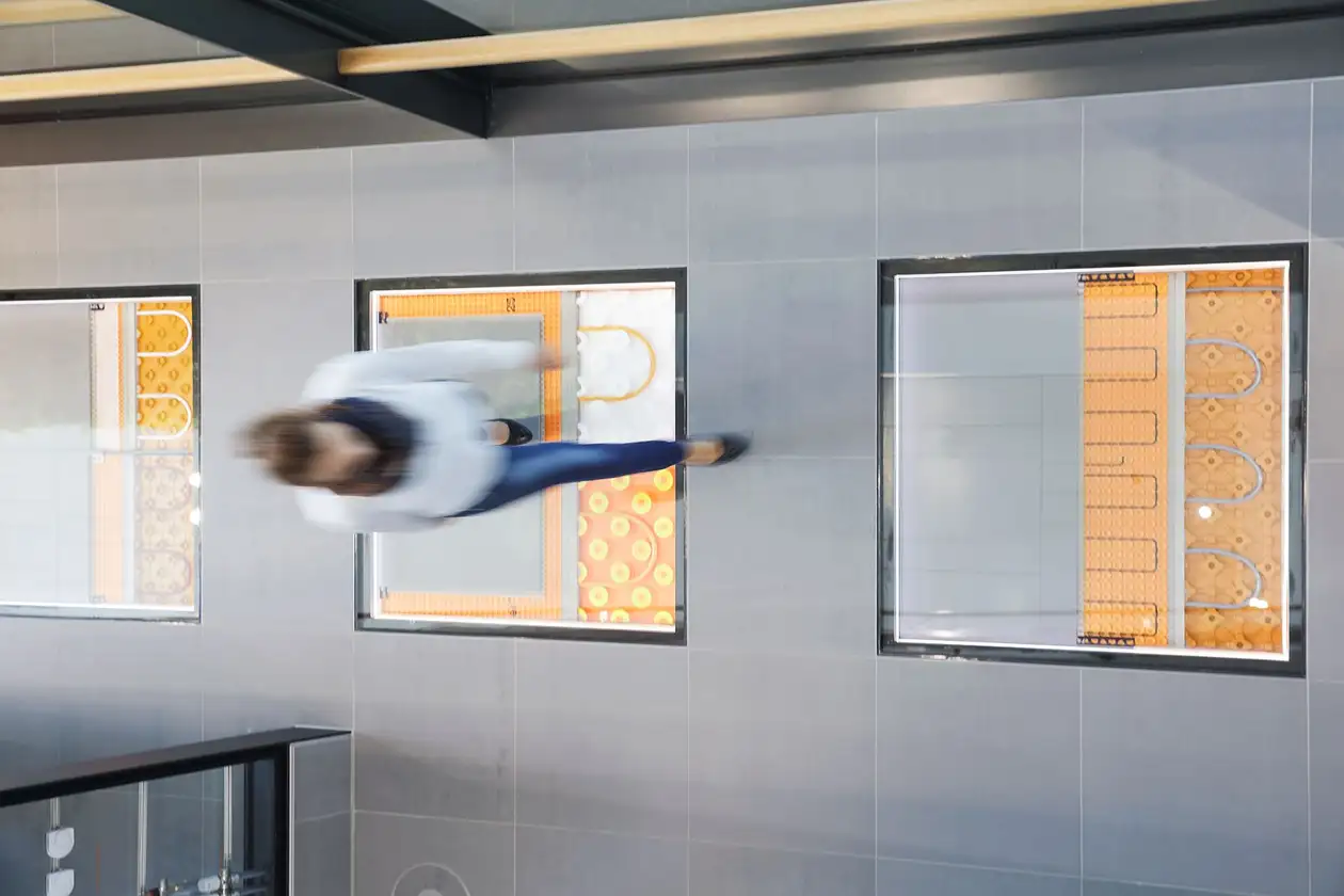 Vue d'en haut sur une femme marchant sur un sol carrelé où des surfaces vitrées laissent entrevoir différents systèmes de chauffage Schlüter-Systems dans le support