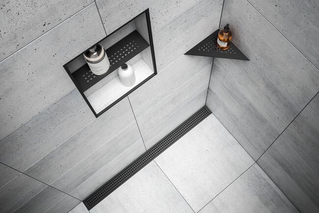 Illustration d'une douche carrelée en aspect béton avec caniveau noir mat Schlüter-KERDI-LINE, tablette de douche de la gamme Schlüter-SHELF et niche avec profilé de finition noir mat.