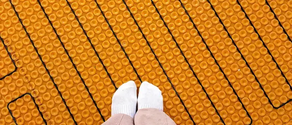 Two women's feet in white socks stand on an orange Schlüter-DITRA-HEAT mat with black heating cables