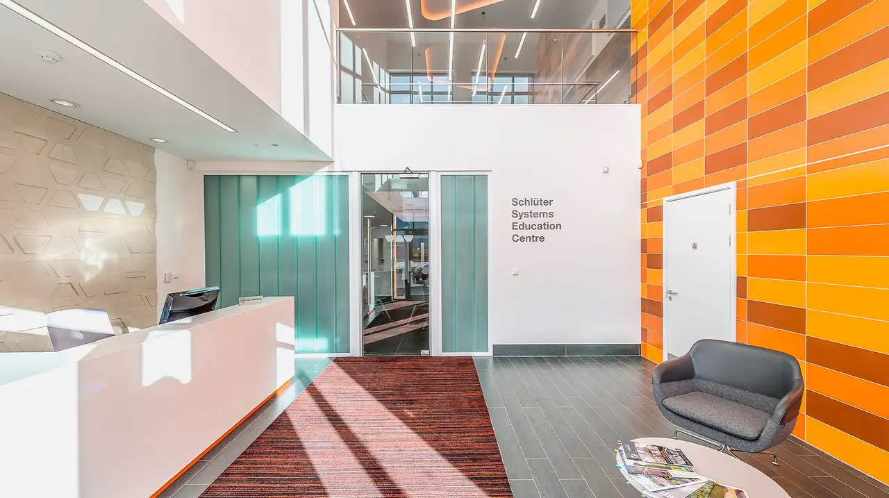 The entrance foyer to Schlüter-Systems Ltd featuring a doorway into the building, a reception desk to the left and seating area on the right. Behind the seating area, a wall with orange tile, heated with DITRA-HEAT