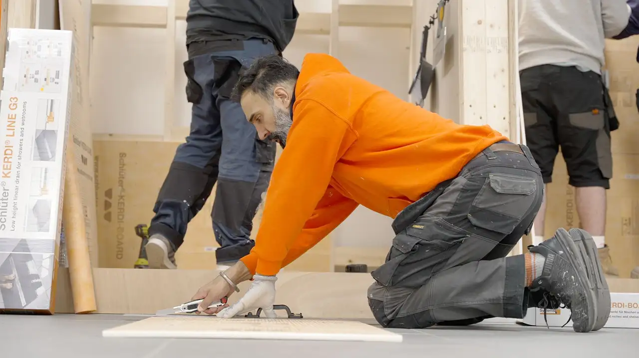 A man in an orange jumper is seen kneeling on the floor and using a utility knife to cut KERDI-BOARD