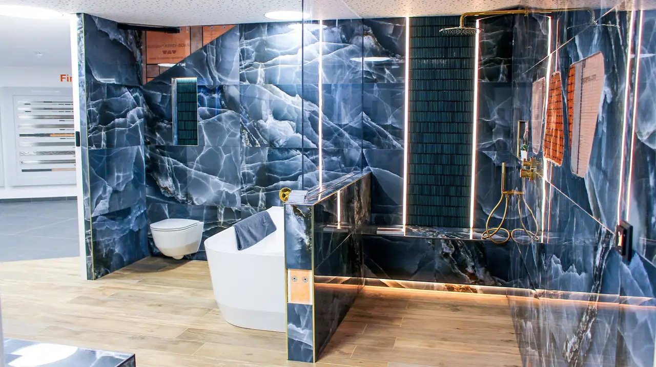A bathroom has been installed in a showroom setting. On the right, a wall hung toilet and a bath are featured, to the left is a walk-in shower. The tile is blue marble and features cutaways to show the Schlüter product