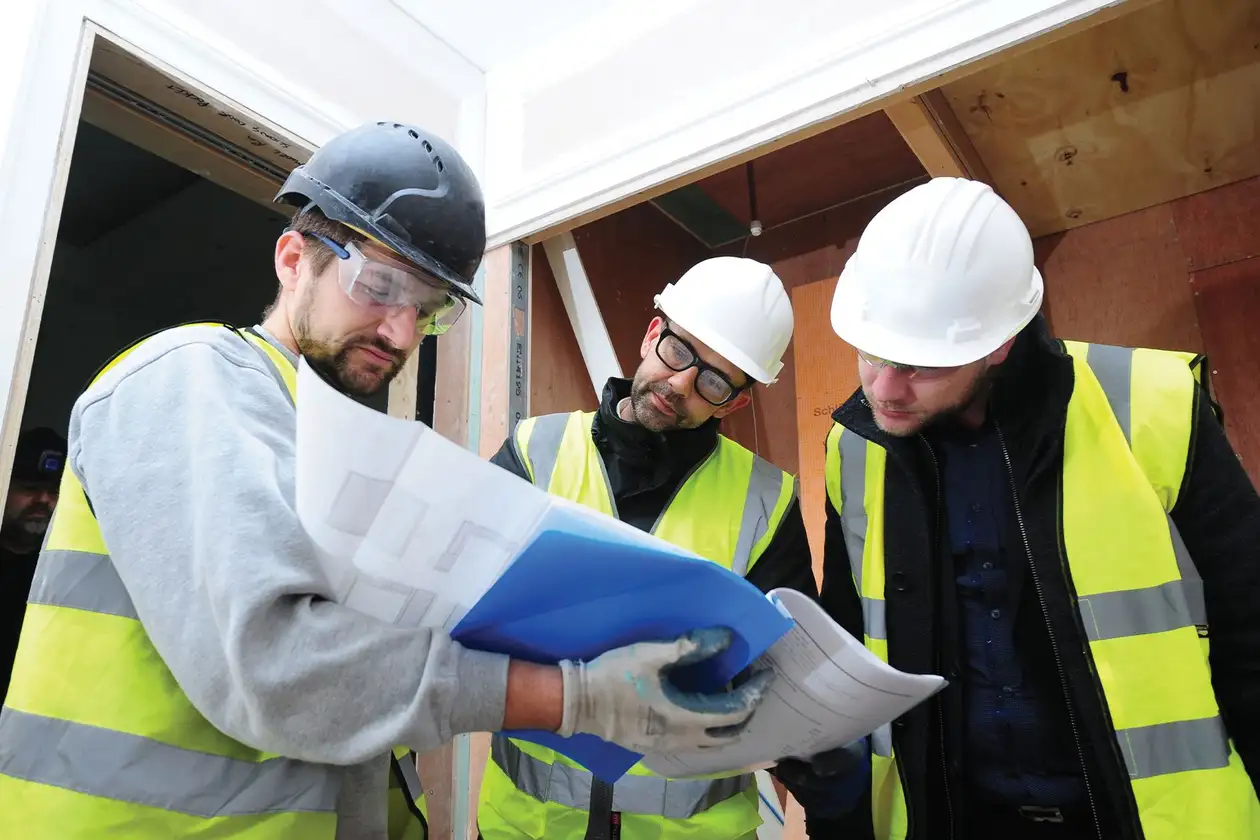 Three people in safety clothing are standing together, looking at the plans for a project