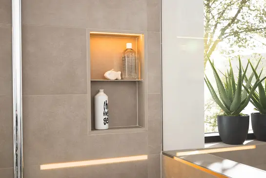 Beige-grey tiled shower wall with a warmly lit, vertical niche for shampoo bottles and toiletries