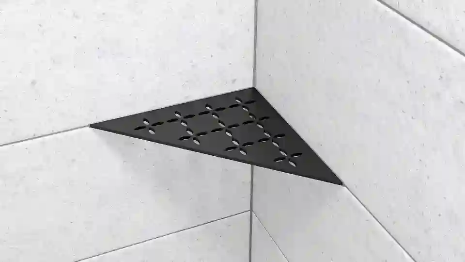Here is a product image of the Schlüter-SHELF-E-S1 shower corner shelf in the shape of an isosceles triangle, in the FLORAL design and MGS matte graphite black finish, installed on light grey wall tiles.