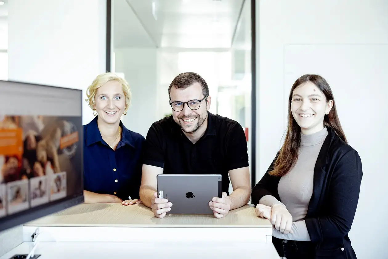 A Schlüter-Systems három munkatársa egy tárgyalóasztalnál áll, a középső táblagépet tart a kezében.