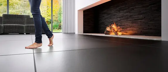 Si vedono le gambe di una donna che cammina a piedi nudi in un soggiorno moderno davanti a un grande camino su un pavimento rivestito con piastrelle scure di grande formato.