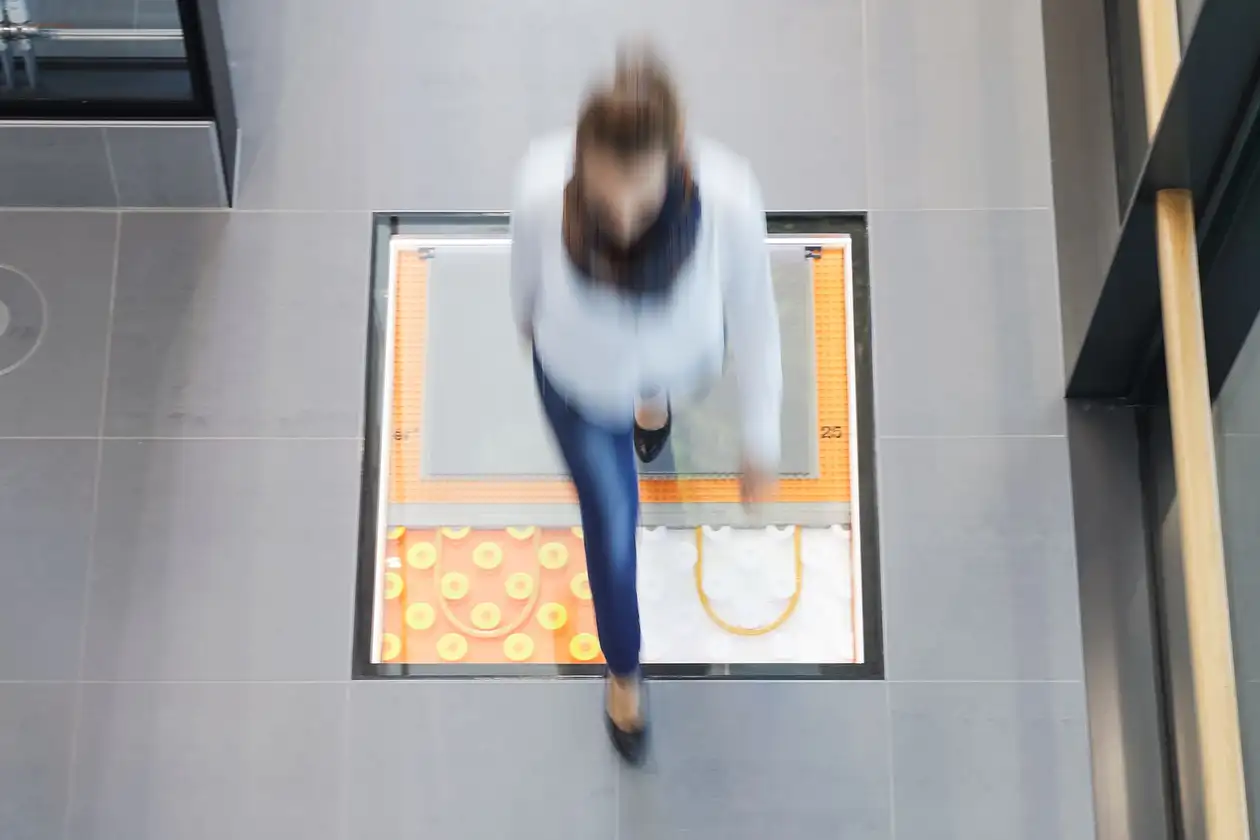 Una donna cammina su un pavimento le cui superfici vetrate offrono una visione dei vari sistemi di riscaldamento Schlüter-Systems presenti nella stratigrafia