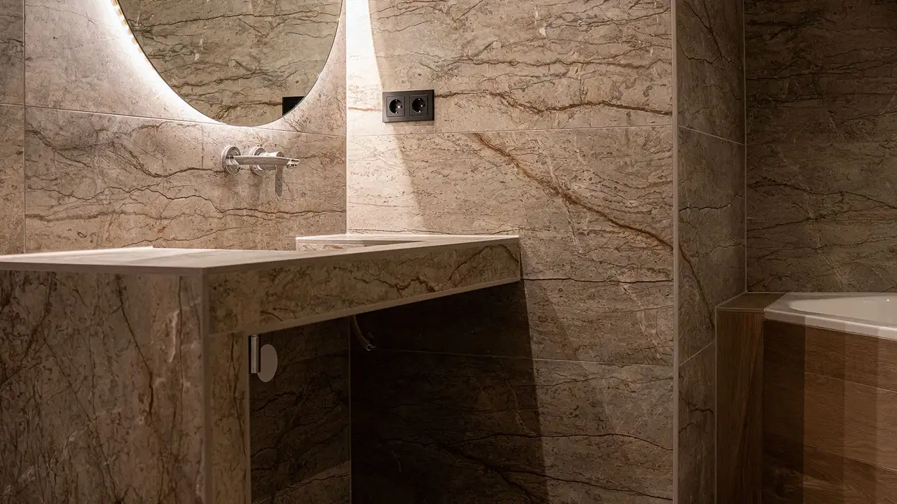 Moderne badkamer met tegels in beige marmerlook, wastafel met Schlüter-JOLLY-afsluitprofiel, verlichte spiegel en zwarte stopcontacten in de wand.