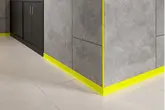Close-up van een grijs betegelde buitenhoek van een wand met gele plint- en wandprofielen