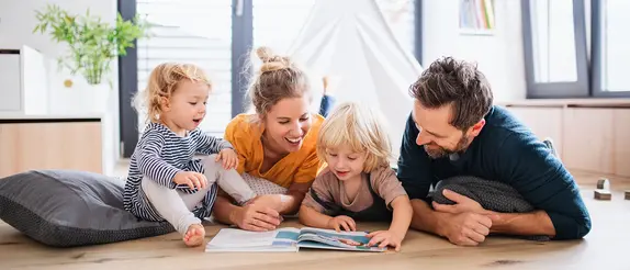 Glad familj best&aring;ende av fem personer, en hund och en katt sitter barfota p&aring; ett gr&aring;tt klinkergolv framf&ouml;r en brun soffa.