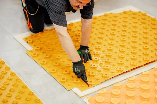 Här syns tre olika orangefärgade BEKOTEC-golvvärmeskivor med noppor som ligger på ett grått kaklat golv. En håller på att skäras till med mattkniv.