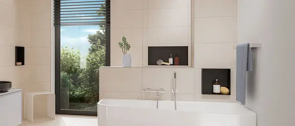 Badrum med ljusbeige platts&auml;ttning, vitt badkar och svarta Schl&uuml;ter-DESIGN-NICHE-v&auml;ggnischer