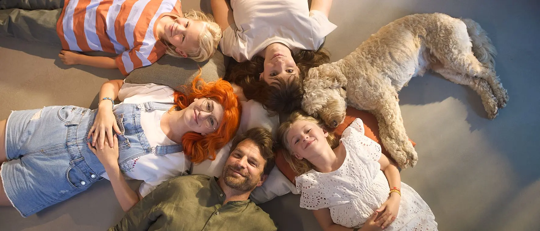 En grupp p&aring; fem personer och en hund ligger p&aring; golvet och tittar leende in i kameran.
