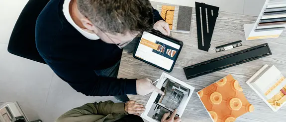 F&aring;gelperspektiv p&aring; en Schl&uuml;ter-anst&auml;lld som pratar med en kund vid ett skrivbord, d&auml;r det finns en rad olika produktprover fr&aring;n Schl&uuml;ter-Systems