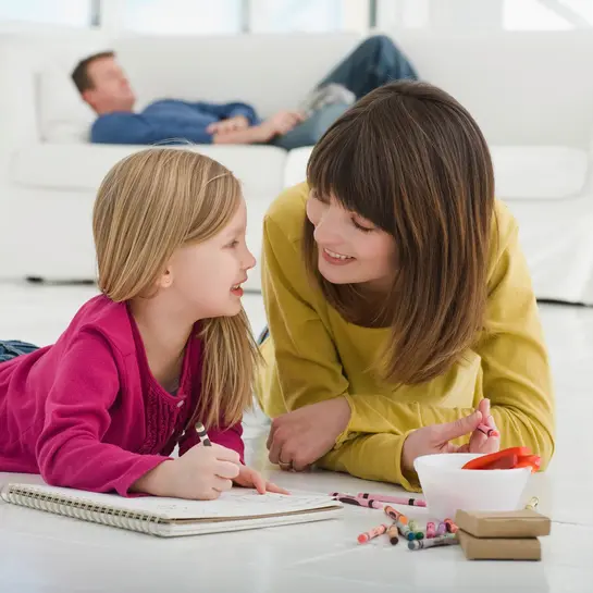 Mamma och dotter ligger framf&ouml;r en vit soffa p&aring; det uppv&auml;rmda klinkergolvet och m&aring;lar.