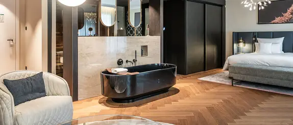 Steigenberger Hof'ta ahşap zemine, d&ouml;şemeli karyolaya ve a&ccedil;ıkta bağımsız duran siyah bir k&uuml;vete sahip modern, sıcak aydınlatılmış otel odası